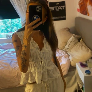 Vit blommig blus med volanger och knyt - Supersöt vit blus med ljusblått blommönster, volangdetaljer och knyt framtill. Ärmlös modell med v-ringning och dubbla volanger i midjan för en romantisk vibe. Perfekt för varma dagar och enkel att styla med jeans eller kjol.