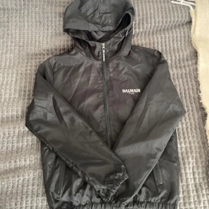 Svart vindjacka från Balmain Paris - Svart vindjacka från Balmain Paris med huva och dragkedja framtill. Jackan har vita Balmain-loggor på bröstet och ryggen, elastiska muddar och två fickor med dragkedja. Perfekt för dig som vill ha en clean och stilren look.