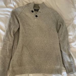 Grå stickad tröja med knappar - Snygg grå stickad tröja från Peacocks med hög krage och två svarta knappar vid halsen. Tröjan har lång ärm, ribbade muddar och en grovstickad framsida som ger en cool textur. Perfekt för kyliga dagar och enkel att matcha med jeans.