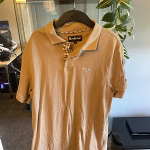 Beige pikétröja från Barbour - Snygg beige pikétröja från Barbour med klassisk krage och korta ärmar. Tröjan har Barbour-logga broderad på bröstet och rutigt tyg vid knäppningen. Perfekt för en clean och stilren look.