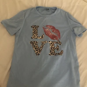 Blå t-shirt med leopardtext från SHEIN - Ljusblå t-shirt från SHEIN med korta ärmar och coolt tryck framtill. Texten LOVE är i leopardmönster och har ett stort rött läppavtryck. Skön passform och mjukt bomullsmaterial, perfekt för en chill look.
