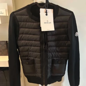 Svart cardigan jacka från Moncler - Snygg svart cardigan jacka från Moncler med quiltad framsida och två fickor med knapp. Jackan har dragkedja och Moncler-logga på ärmen. Perfekt för dig som vill ha en stilren och sportig look.
