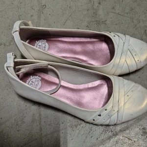 Ballerinaskor - Snygga beige guld ballerinaskor från Marmelata med rund tå, dekorativa remmar och små silvriga nitar framtill. Skorna har en smal rem runt vristen och en mjuk, ljusrosa innersula som ger en feminin touch. Perfekta för dig som gillar stilrena och söta skor.