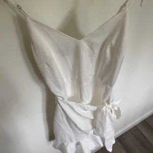 Vit kort omlottklänning med volang - Supersöt vit kort klänning med smala axelband, omlottknytning i midjan och volang nertill. Perfekt för sommarens alla tillfällen. Klänningen har en lätt v-ringning och är tillverkad i ett tunt, mjukt material.