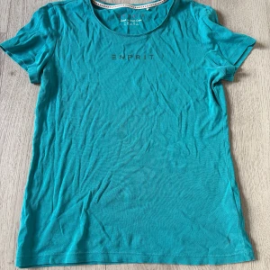 Turkos t-shirt från Esprit med strass - Turkos t-shirt från Esprit i mjuk bomull med korta ärmar och rund halsringning. Framsidan har Esprit-loggan i glittriga strassdetaljer som ger en snygg touch. Perfekt till jeans eller shorts för en chill look.