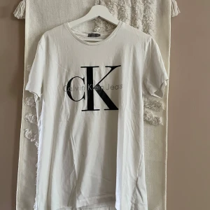 Vit t-shirt från Calvin Klein Jeans - Vit t-shirt från Calvin Klein Jeans med stor svart CK-logga framtill. Klassisk rund halsringning och korta ärmar. Tillverkad i 100% bomull för en skön och luftig känsla. Perfekt för en clean och stilren look.