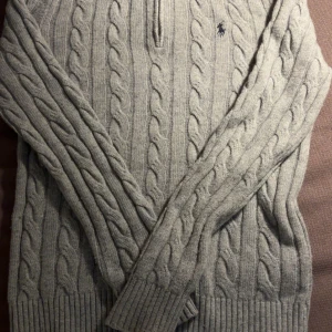 Grå kabelstickad tröja Polo Ralph Lauren - Kabelstickad grå tröja från Polo Ralph Lauren med halv dragkedja och klassisk broderad logga på bröstet. Tröjan har ribbade muddar, hög krage och är långärmad. Perfekt för dig som gillar stilren och tidlös design.
