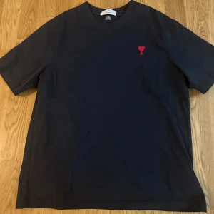 Svart ami paris t-shirt - Svart t-shirt från Ami med klassisk passform och korta ärmar. På bröstet finns det ikoniska broderade röda hjärtat med bokstaven A. Tillverkad i mjuk bomull som känns skön mot huden. Perfekt för en clean och stilren look.