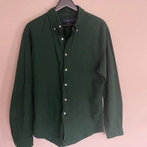 Grön skjorta från Ralph Lauren - Snygg grön skjorta från Ralph Lauren med klassisk button-down krage och vita knappar. Skjortan har lång ärm och den ikoniska lilla broderade loggan på bröstet. Perfekt för dig som gillar stilrena och tidlösa plagg.