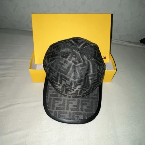 Svart Fendi keps med FF-mönster - Snygg svart keps från Fendi med ikoniskt FF-monogram över hela kepsen. Klassisk böjd skärm och justerbar rem bak med metallogo. Tillverkad i bomullsmix och levereras med original Fendi-box och tagg. Perfekt för dig som vill sticka ut med lyxig streetstyle.
