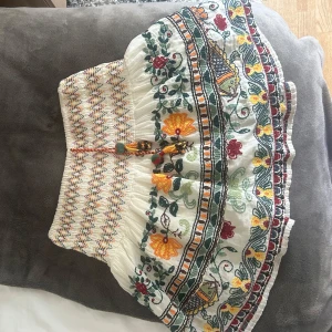 Zara kjol - Superfin vit kortkjol med broderade blommor i gult, rött och grönt. Kjolen har elastisk midja, snörning med tofsar och små träpärlor. Går inte att köpa längre och superunik! 