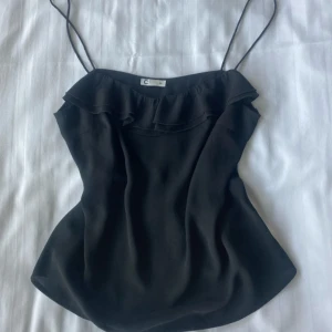 Svart linnetopp med volang  - Svart linnetopp med tunna axelband och snygg volangdetalj längs urringningen. Toppen har en lätt utsvängd passform och är tillverkad i ett mjukt, slätt tyg som ger en stilren look. 💖☺️