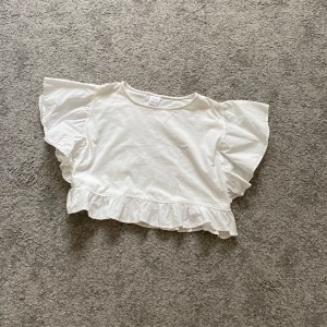 Vit volangblus från Zara - Supersöt vit blus från Zara med breda volangärmar och volangkant nertill. Den har en luftig och croppad passform som känns riktigt trendig. Perfekt till jeans eller kjol för en fräsch look. Gjord i mjuk bomull som är skön mot huden.