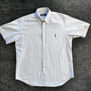Ralph Lauren Skjorta  - Kortärmad skjorta från Polo Ralph Lauren i ljusgrå och vit tunn rand. Klassisk krage, knappstängning och bröstficka med broderad logga. Skjortan är i 100% bomull och har en clean, tidlös look som funkar till allt. Skick 10/10 som ny, Storlek L. Pris går att diskutera✅