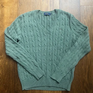 Grön kabelstickad tröja Polo Ralph Lauren - Kabelstickad grön tröja från Polo Ralph Lauren med v-ringning och broderad logga på bröstet. Tröjan har långa ärmar, ribbade muddar och är gjord i mjuk bomull. Perfekt för dig som gillar klassisk och stilren look. Kids size M. Längd 55 cm, bredd 45 cm. Fraktar oftast samma dag!!😁