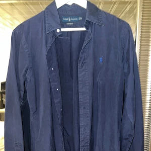 Mörkblå skjorta från Ralph Lauren - Klassisk mörkblå skjorta från Ralph Lauren med broderad logga på bröstet. Skjortan har lång ärm, knappar framtill och en snygg krage. Perfekt för en stilren och avslappnad look.