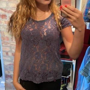 Lila spetsblus med kort ärm - Säljer en lila blus i transparent spets med blommigt mönster. Toppen har rund halsringning och korta ärmar, perfekt för dig som vill ha en cool och trendig look. Superfin att styla lager på lager eller med ett linne under.