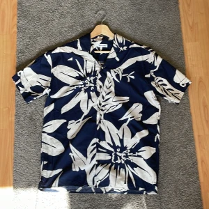 Blommig skjorta från Jack & Jones - Snygg kortärmad skjorta från Jack & Jones i marinblått med stora vita blommor. Skjortan har klassisk krage, knäppning framtill och är tillverkad i mjuk bomull. Perfekt för sommarens alla äventyr.