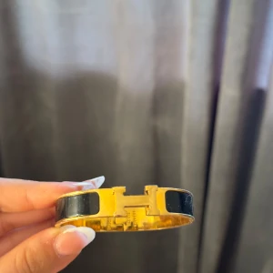 Svart och guld H- armband - Snyggt armband  i guldtonad metall med svarta detaljer och ikoniskt H-spänne framtill. Perfekt statement piece för dig som gillar lyxiga accessoarer med klassisk design. Armbandet har en stilren och tidlös look som lyfter vilken outfit som helst.