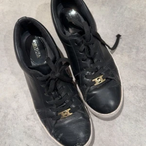 Svarta sneakers från Michael Kors - Stilrena svarta sneakers från Michael Kors i skinn med vita sulor och svarta snören. Snygg guldfärgad MK-detalj framtill och logga på sidan av sulan. Perfekta för dig som gillar klassisk streetstyle med en touch av lyx. Använda som man ser, men fortfarande användbara. Köpta för 1800