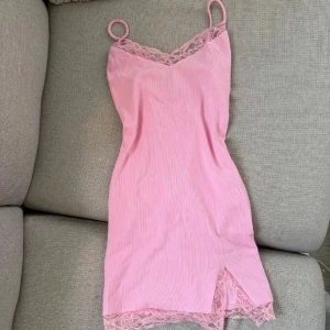 Rosa klänning med spetsdetaljer - Söt rosa miniklänning med smala axelband och spetsdetaljer vid halsringning och nederkant. Klänningen har en liten slits framtill och är figurnära med en mjuk, ribbad känsla. Storlek s men passar mig som brukar ha xs