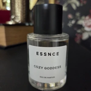 ESSNCE Cozy Goddess Eau de Parfum - Parfymen Cozy Goddess från ESSNCE kommer i en stilren, rund glasflaska med svart lock. Doften har tydliga inslag av vanilj, varm krydda, pudriga och träiga toner samt balsamiska nyanser. Noter av kastanj, vanilj, peppar, sandelträ, äpple och cashmeran. SE INNEHÅLL PÅ BILD.