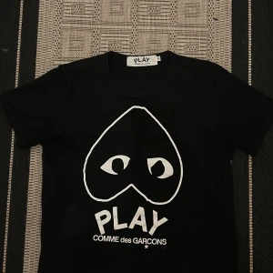 Svart t-shirt Comme des Garçons Play - Svart t-shirt från Comme des Garçons Play med ikoniskt hjärta med ögon tryckt i vitt på bröstet. Klassisk rund hals och korta ärmar. Skön och stilren, perfekt för dig som gillar streetwear och vill sticka ut med en grafisk look.