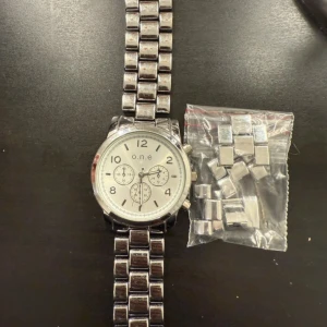 Silverfärgad klocka från o.n.e - Stilren klocka från o.n.e med silverfärgad länkarmband och rund urtavla. Urtavlan är ljus med tydliga siffror och tre mindre visartavlor. Klockan levereras i originalask och extra länkar medföljer för enkel justering av storlek. Lite missfärgad som ni kan se på bild, behöver batteribyte 