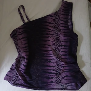 Lila oneshoulder topp med mönster - Snygg lila oneshoulder topp med mörklila och svarta vågiga och prickiga mönster. Tunt och lätt material. En axelbandslös sida och ett smalt axelband på andra sidan ger en cool asymmetrisk look.