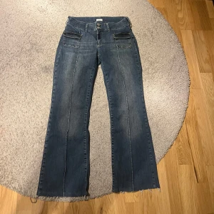 Blå bootcut jeans från Nelly - Snygga blå jeans från Nelly med bootcut passform och råa, fransiga benslut. Jeansen har två framfickor med lock och markerade sömmar längs benen. Klassisk femficksmodell och dubbla knappar i midjan. Perfekta för en trendig vardagslook.
