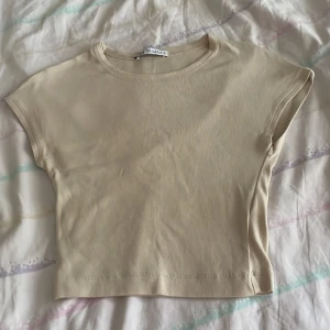 Beige croppad topp från Zara - Enkel och stilren croppad topp från Zara i beige färg. T-shirten har korta ärmar och rund halsringning. Perfekt att matcha med jeans eller kjol för en clean look. Mjuk och skön bomullskänsla.