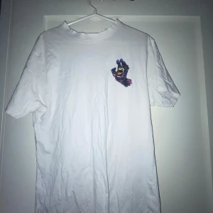 Vit Santa Cruz t-shirt med färggrafik - Vit t-shirt från Santa Cruz med en stor, färgstark grafik av en skrikande hand på ryggen och en mindre version på bröstet. T-shirten är i bomull och har klassisk passform med korta ärmar. Perfekt för dig som gillar streetwear och unika prints.