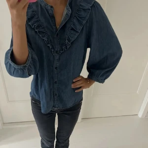 Jeansskjorta/blus med volang från Holly & Whyte - Snygg jeansskjorta/blus från Holly & Whyte med volangdetaljer längs bröstet och axlarna. Skjortan har långa ärmar med resår vid ärmslut och knäppning framtill. Perfekt för dig som gillar en avslappnad men ändå trendig look. Nypris 500kr.🩵