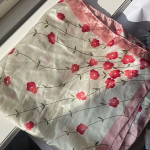 Blommig ljusrosa sidenscarf - Superfin fyrkantig scarf i glansigt sidenliknande material. Ljusrosa kant och blommigt mönster med röda och rosa blommor på vit botten. Perfekt att knyta runt halsen eller i håret för en söt och fräsch look.