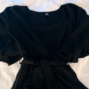 Svart omlottklänning med knyte - Supersnygg svart omlottblus med knytband i midjan och vida korta ärmar. Blusen har en flowy känsla och är perfekt för dig som gillar en enkel men chic stil. Passar lika bra med sneakers som med klackar.