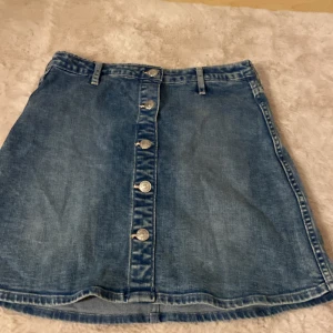 Jeanskjol med knappar från H&M - Snygg jeanskjol från H&M i klassisk blå denim. Kjolen är kort och har en rak passform med knappar framtill och hällor för bälte. Perfekt till sneakers eller boots för en chill look.