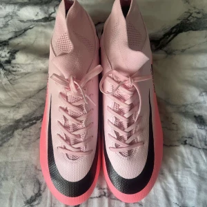 Rosa Nike fotbollsskor med snörning - Snygga fotbollsskor från Nike i ljusrosa med svart swoosh längs sidan. Skorna har högre skaft, snörning och är tillverkade i syntetmaterial med ventilerande meshdetaljer. Perfekta för dig som vill sticka ut på planen.