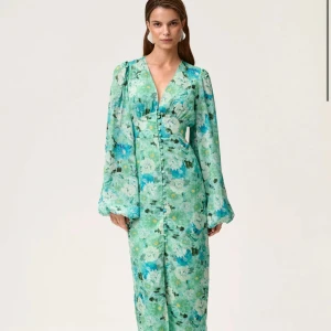 Grön blommig långklänning med v-ringning - Superfin långklänning i grönt med blommigt mönster och v-ringad hals. Klänningen har långa, luftiga ärmar och knappar framtill. Perfekt för dig som vill sticka ut med en färgglad och romantisk look. Använd endast en kväll så i nyskick. 