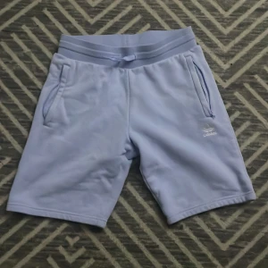 Ljuslila Adidas shorts med snörning - Snygga ljuslila shorts från Adidas med elastisk midja och snörning. Mjukt material i polyester, två fickor med dragkedja och diskret Adidas-logga framtill. Perfekta för chill dagar eller träning. PRIS KAN DISKUTERAS 