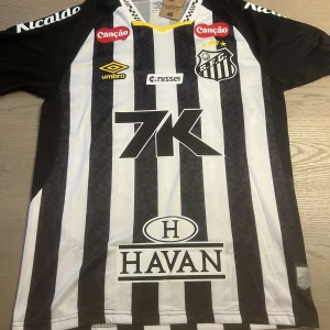 Santos FC Neymar Jr. matchtröja Umbro - Santos FC fotbollströja med Neymar Jr. och nummer 10 på ryggen. Tröjan är svart och vit med vertikala ränder, flera sponsorloggor och klubbmärke på bröstet. Tillverkad i lätt och ventilerande material, perfekt för dig som älskar brasiliansk fotboll.