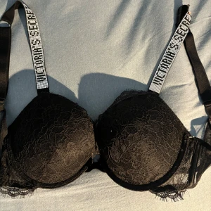 Svart spets-bh Victoria's Secret - Push-up bh från Victoria's Secret i svart spets med vadderade kupor och snygga axelband med strass och logga. Axelbanden är justerbara och bh:n har fina spetsdetaljer längs kanterna. Perfekt för dig som vill ha något extra under outfiten.