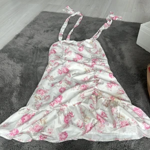 Vit blommig kortklänning från SHEIN - Supersöt vit kortklänning från SHEIN med rosa blommigt mönster. Klänningen har knytbara axelband, volang nertill och dragkedja i sidan. Materialet är glansigt och känns lyxigt, perfekt för sommarens alla tillfällen. Sitter figurnära och har romantiska vibbar.