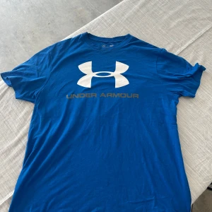Blå Under Armour t-shirt loose fit - Blå t-shirt från Under Armour med stort vitt logotryck på bröstet och texten UNDER ARMOUR under. Modellen är loose fit och tillverkad i ett mjukt bomullsmaterial. Perfekt för dig som gillar sportig och avslappnad stil.