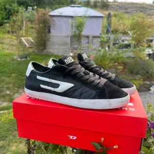 Svarta sneakers från Diesel - Snygga svarta sneakers från Diesel med vit sula och vita detaljer. Skorna har snörning och är tillverkade i skinn och mocka. Perfekta för en stilren look. Liten spricka i sulan bara.
