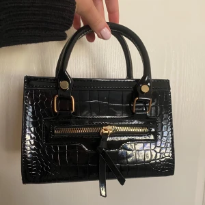 Svart handväska med krokodilmönster - Snygg svart liten handväska med krokodilmönster och guldfärgade detaljer. Väskan har en dragkedja framtill och två handtag. Aldrig använd🥰