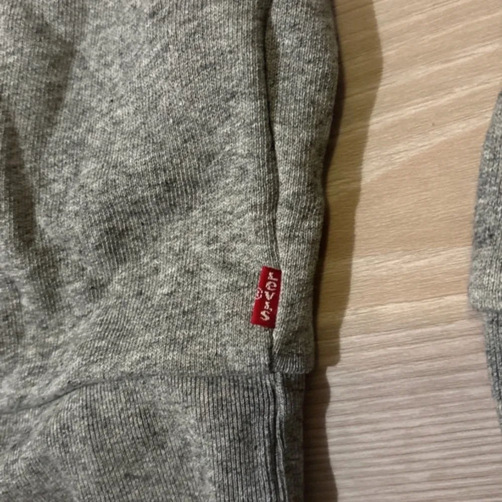 Säljer en grå sweatshirt från Levi's med deras ikoniska röda logga på bröstet. Tröjan har långa ärmar och en rund halsringning. Perfekt för en avslappnad stil. 🩶. Neuletakit & Villapaidat.