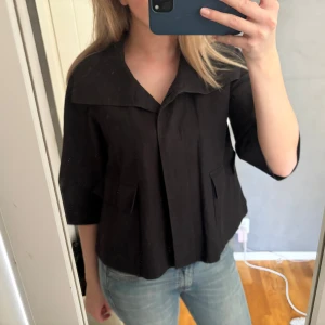 Svart jacka  - Snygg svart jacka med trekvartsärm och stor krage. Jackan har en stilren design med dolda knappar och två fickor framtill. Märket är Taifun.