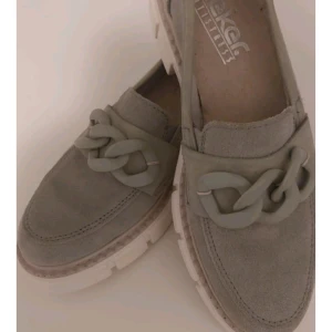 Grå loafers med kedjedetalj - Snygga grå loafers från Rieker med en cool kedjedetalj på ovansidan. Skorna har en robust vit sula som ger en modern touch. Perfekta för en stilren look.