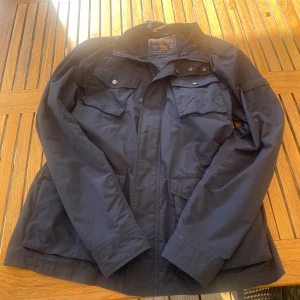 Blå field jacka från Woolrich - Säljer en blå field jacka från Woolrich med flera praktiska fickor och dragkedja. Jackan har långa ärmar och knappar på fickorna. Perfekt för höst och vår. Den är storlek M/L, skulle säga att den är typ i mitten 