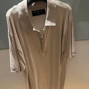 Beige pikétröja från Daniel De Prato - Snygg beige pikétröja från Daniel De Prato med dragkedja framtill. Tröjan har korta ärmar och en klassisk krage, perfekt för en stilren look. Tillverkad i Italien.